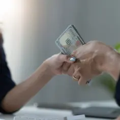 Dwie dłonie przekazują sobie plik banknotów. Jedna z nich ma złoty pierścionek. To symboliczna scena zawarcia umowy pożyczki.