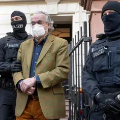 Mężczyzna w garniturze i masce, zakuty w kajdanki, prowadzony przez policjantów. Wygląda jak scena z filmu o próbie puczu.