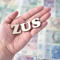 Litery "ZUS" na dłoni na tle polskich banknotów. Symbolizuje to potencjalne odszkodowanie z ZUS.