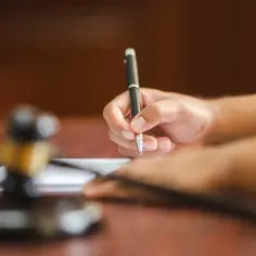 Dłoń referendarza sądowego pisze piórem na papierze, obok leży młotek sędziowski.