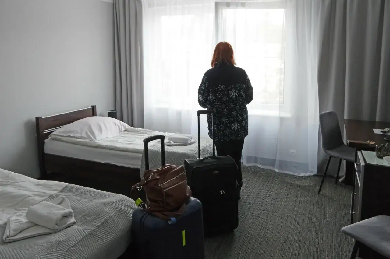 Kobieta z walizkami w pokoju hotelowym, wyglądającym jak najgorsze sanatorium z ZUS.