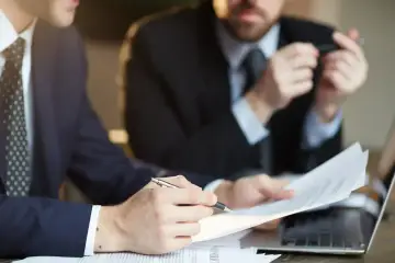 Dwóch mężczyzn w garniturach analizuje dokumenty. Jeden z nich pisze piórem, drugi trzyma długopis. Nagana w pracy? Nie, to poważna rozmowa o kontrakcie.